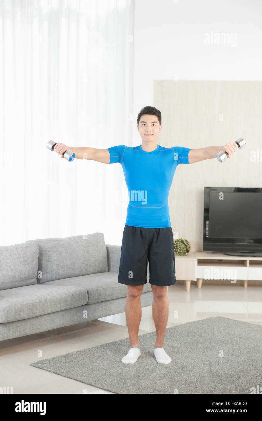 Man standing and opening arms with dumbbells staring at front in living room Stock Photo