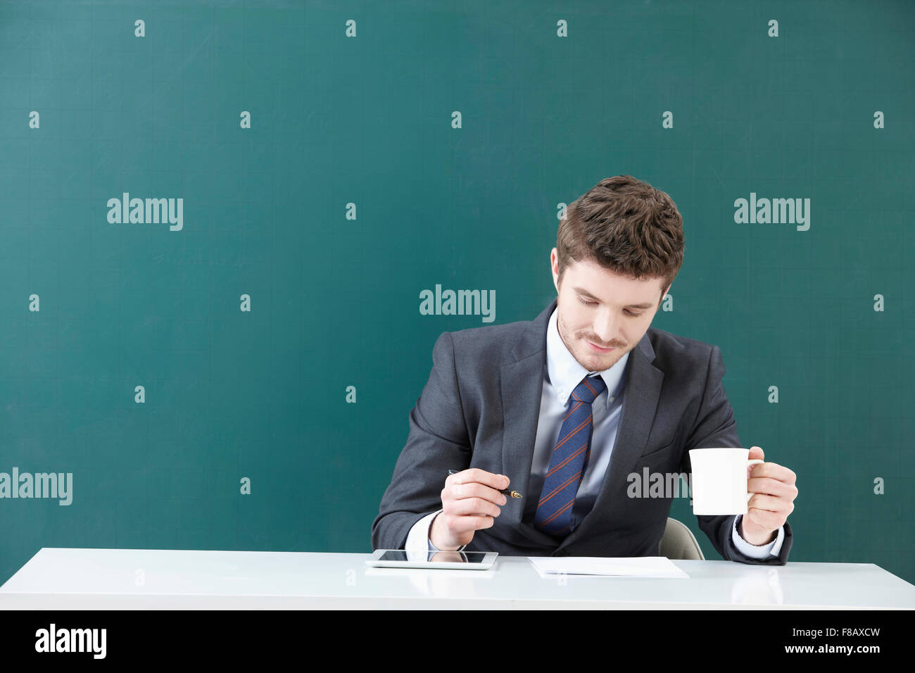 Portrait of man in suit holding a mug and a pen with tablet and paper on table in front of blackboard Stock Photo