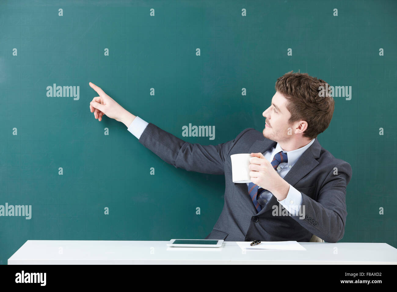 Portrait of man in suit pointing at blackboard with a mug in his hand and tablet, pen, and paper on the table Stock Photo
