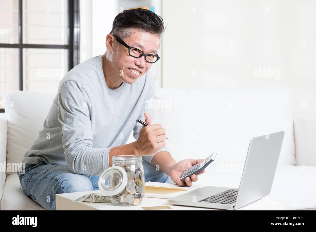 Mature 50s Asian man giving thumb up while counting on money using calculator and laptop