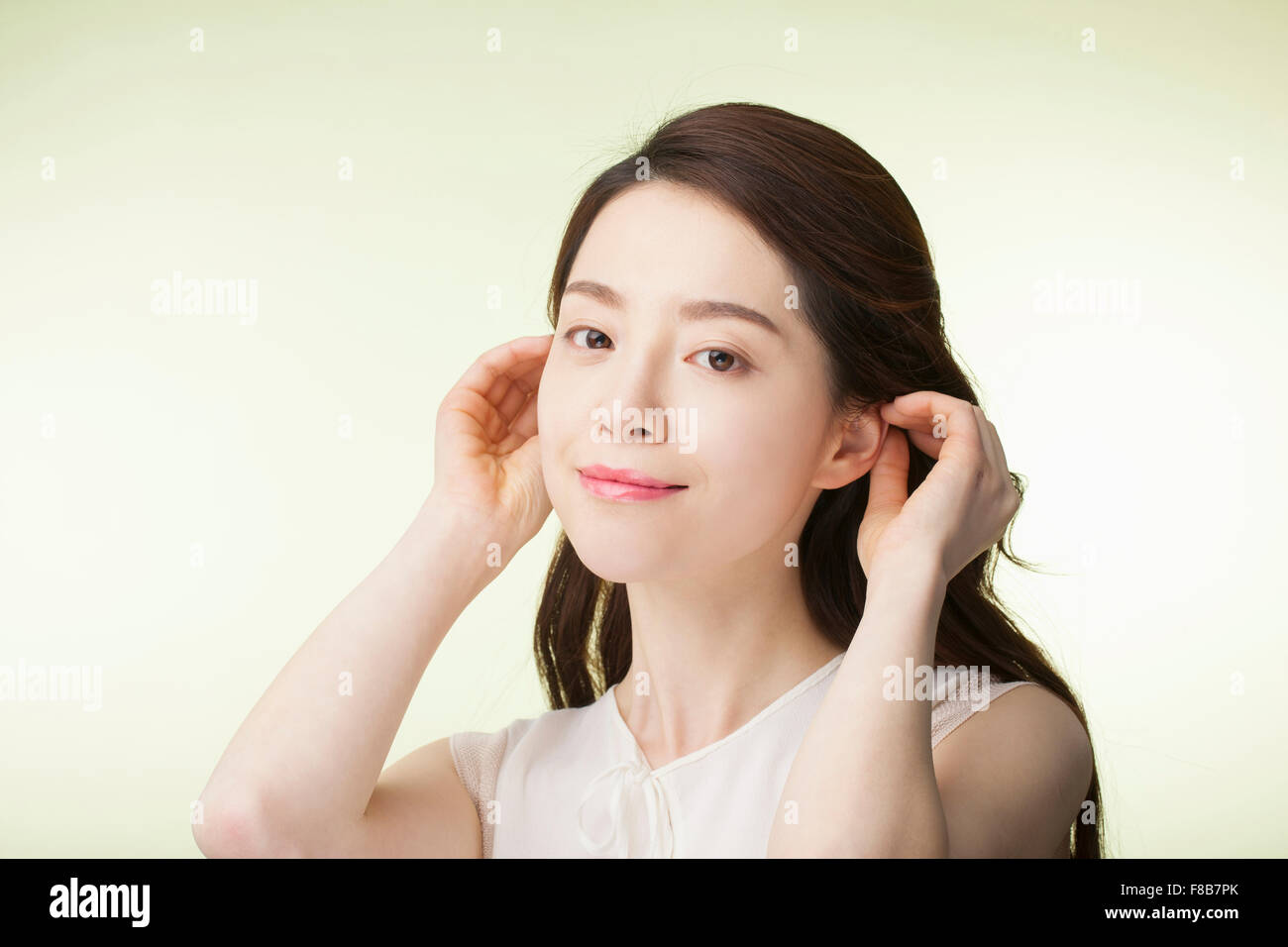 Korean woman with long wavy hair in white sleeveless shirt touching her ears and smiling Stock Photo