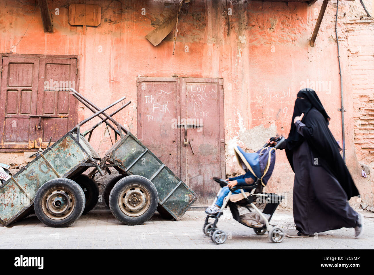 Street photography. Muslim woman wearing a black burqa pushing a push chair past some rusty carts Stock Photo