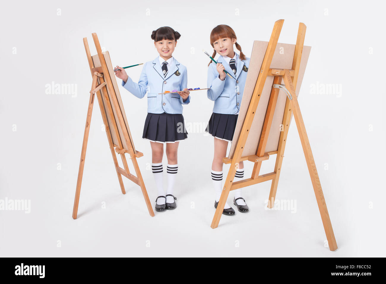 Two elementary school girls in school uniforms standing with easels and holding brushes both staring forward with a smile Stock Photo