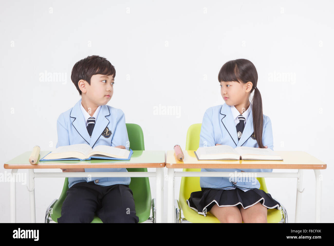 Elementary school boy and girl in school uniforms seated at desk with big pencils and books on their desk and looking at each Stock Photo