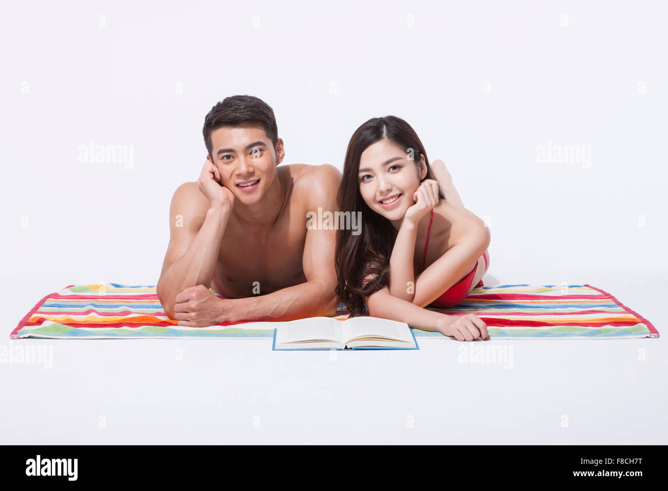 Couple in beach wear lying on front on a colorful towel with a book and staring forward with a smile with their hand under Stock Photo