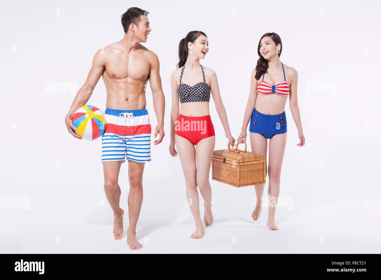 Man in swimming pants holding a beach ball and two women holding a picnic basket together all walking together Stock Photo