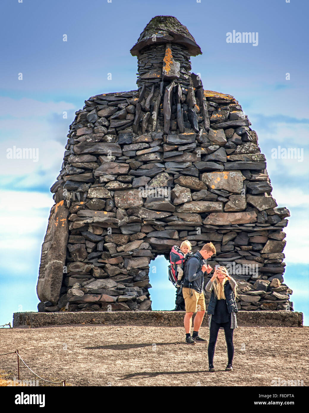 Arnarstapi, Snaefellsnes Peninsula, Iceland. 30th July, 2015. A tourist with a child in a backpack carrier, and his companion looking at her smartphone, visit the huge stone structure of Bardur SnaefellsÃ¡s at Arnarstapi on the Snaefellsnes peninsula in West Iceland. By sculptor Ragnar Kjartansson, it relates to a late Icelander saga with legendary elements dating to the early 14th century. It is a favorite tourist destination. © Arnold Drapkin/ZUMA Wire/Alamy Live News Stock Photo