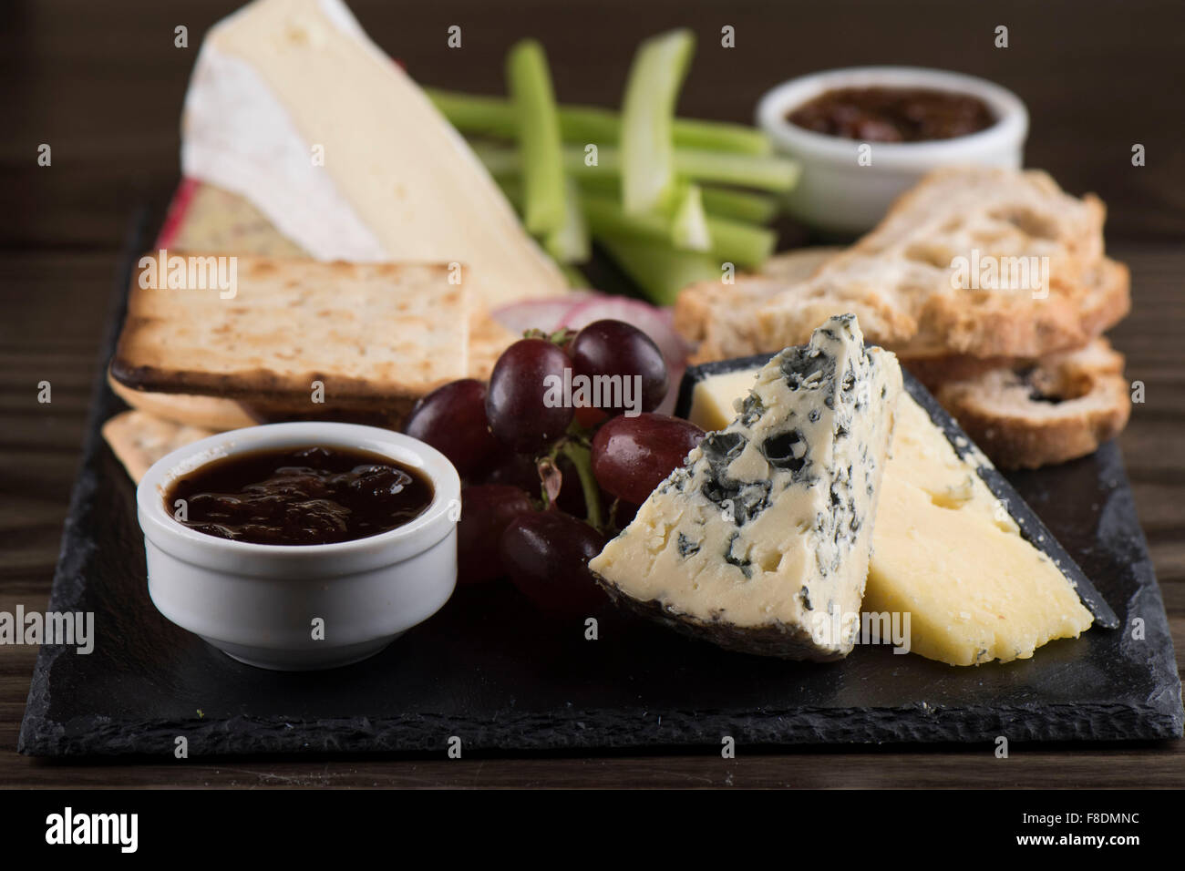 A cheese plate on slate with chutney, cheddar, stilton cheese, crackers, brie, grapes and celery. Stock Photo