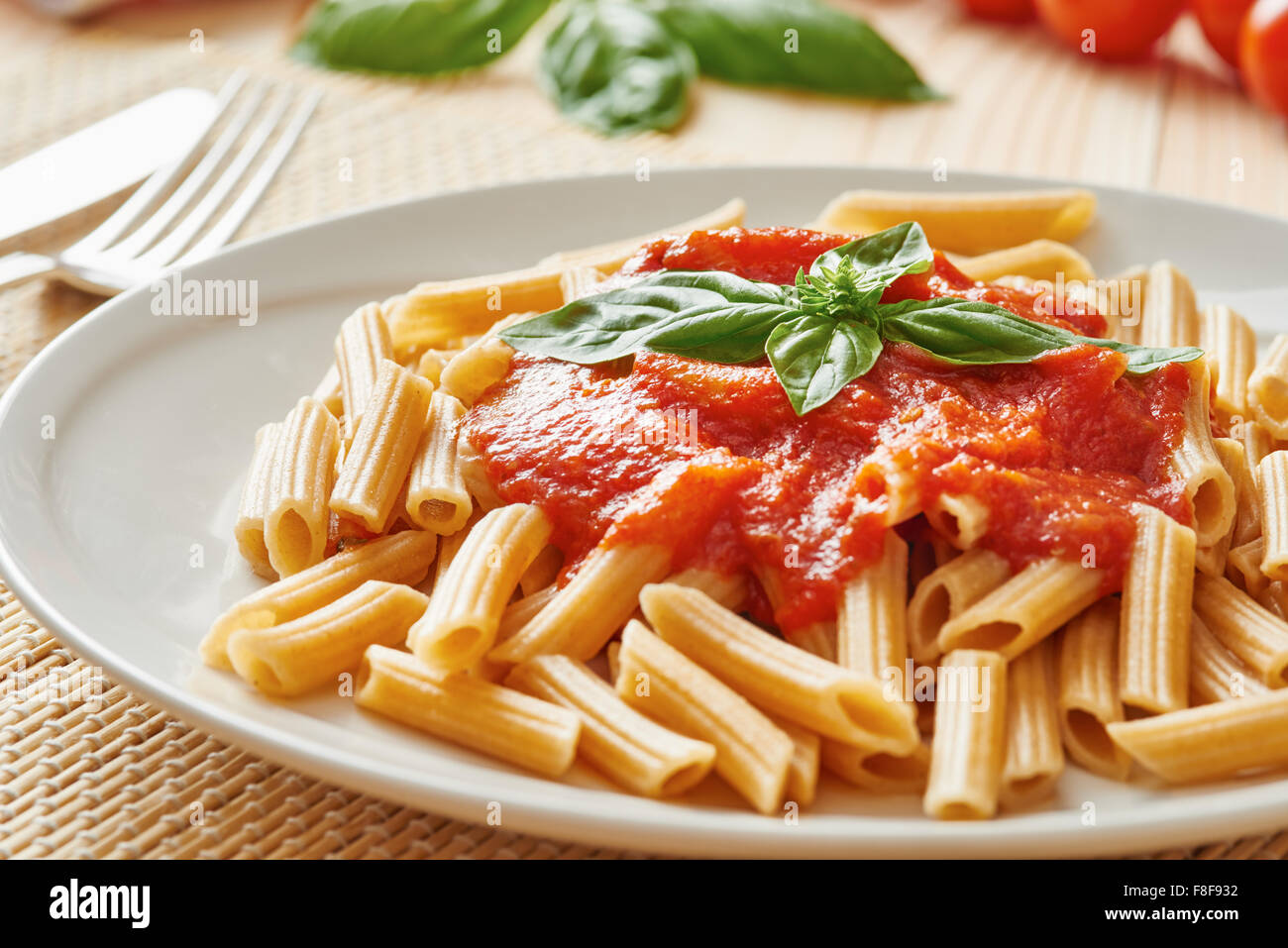 Wheat pasta with tomato sauce on white plate surrounded by basil and tomato on wooden table Stock Photo
