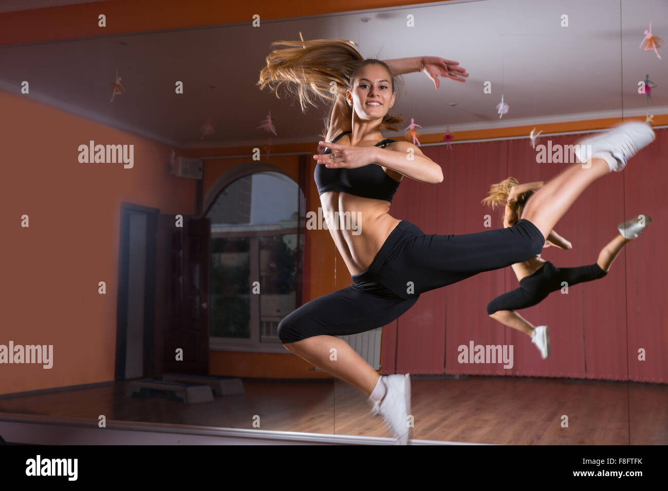 Full Length Profile of Young Blond Female Dancer Leaping in Air and Smiling at Camera in Dance Studio in front of Mirrored Wall Stock Photo