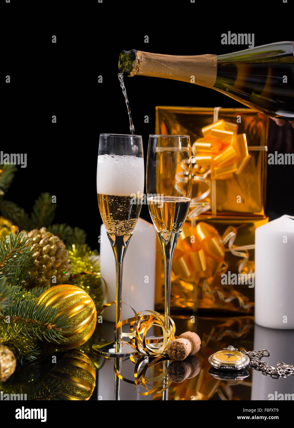 Festive Still Life - Pouring Champagne into Glasses in front of Black Background with White Candles, Gold Wrapped Gifts and Evergreen Decorated with Christmas Balls and Tinsel. Stock Photo