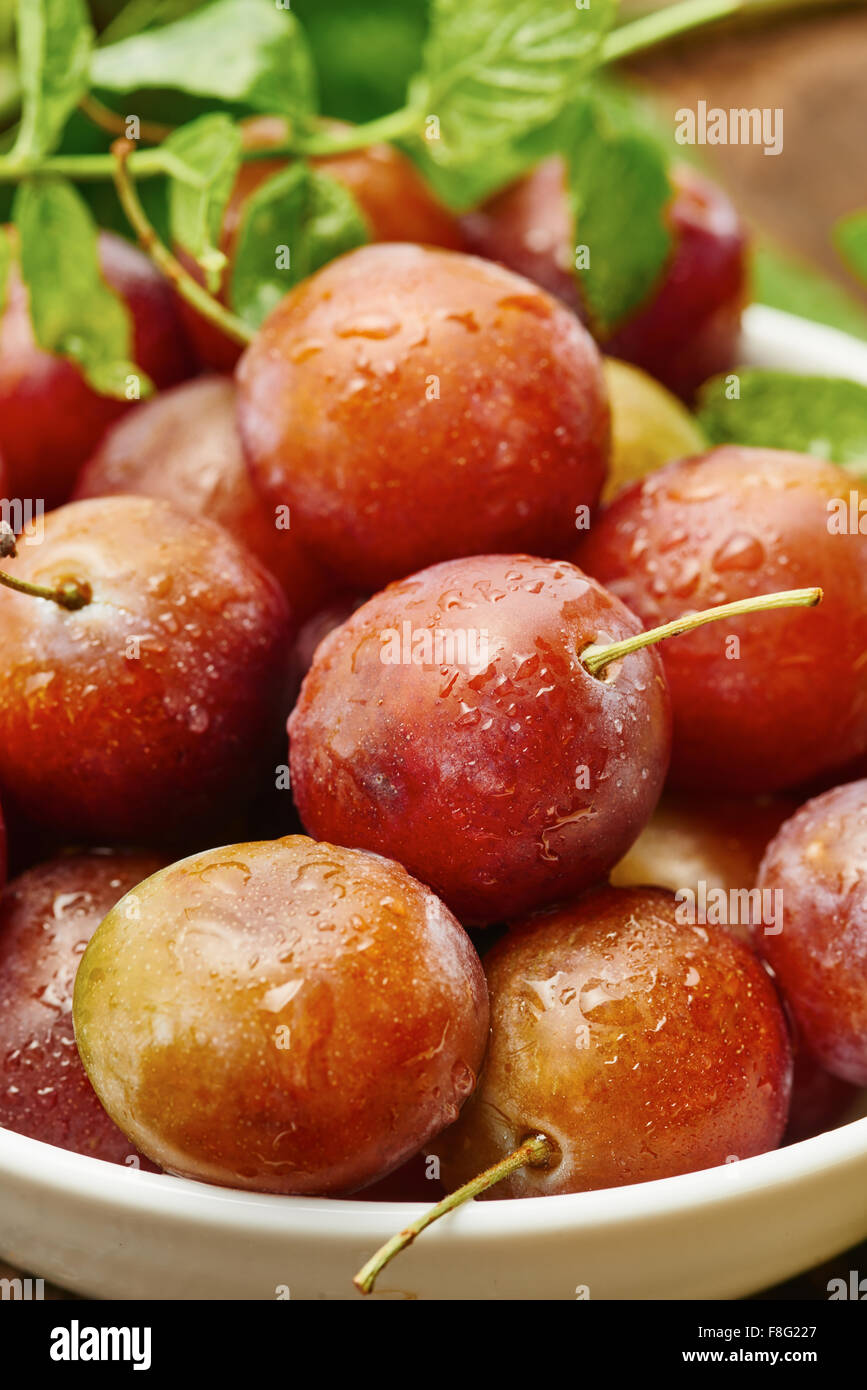 Red plums in bowl on wood surrounded by lemon and green Stock Photo