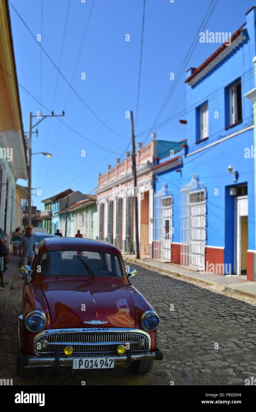 vintage dark red Hillman car parked in the shade and pedestrians walking down a colorful cobbled street in Trinidad Cuba Stock Photo