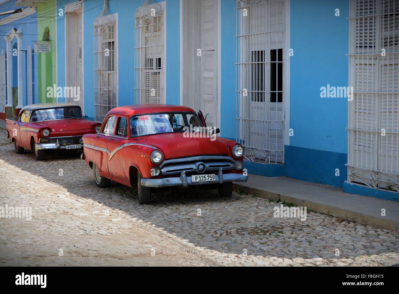 two red vintage cars parked up in the shade on a cobbled street lined with pastel colored buildings in Trinidad Cuba Stock Photo