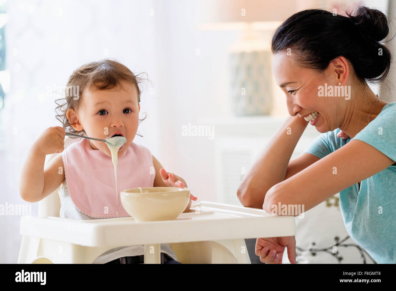 Mother watching baby daughter eat in high chair Stock Photo