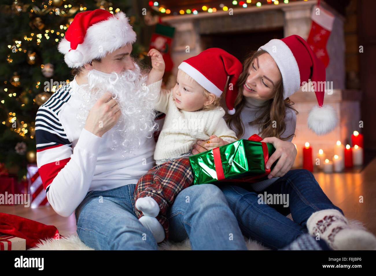 Surprised child looks at dad weared Santa Claus with fake beard sitting opposite Christmas tree Stock Photo