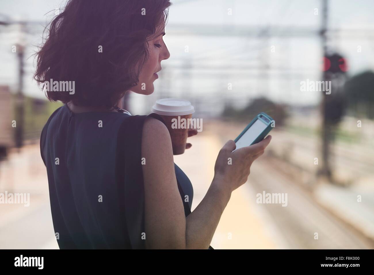Mid adult woman waiting at train station, using smartphone, holding coffee cup Stock Photo