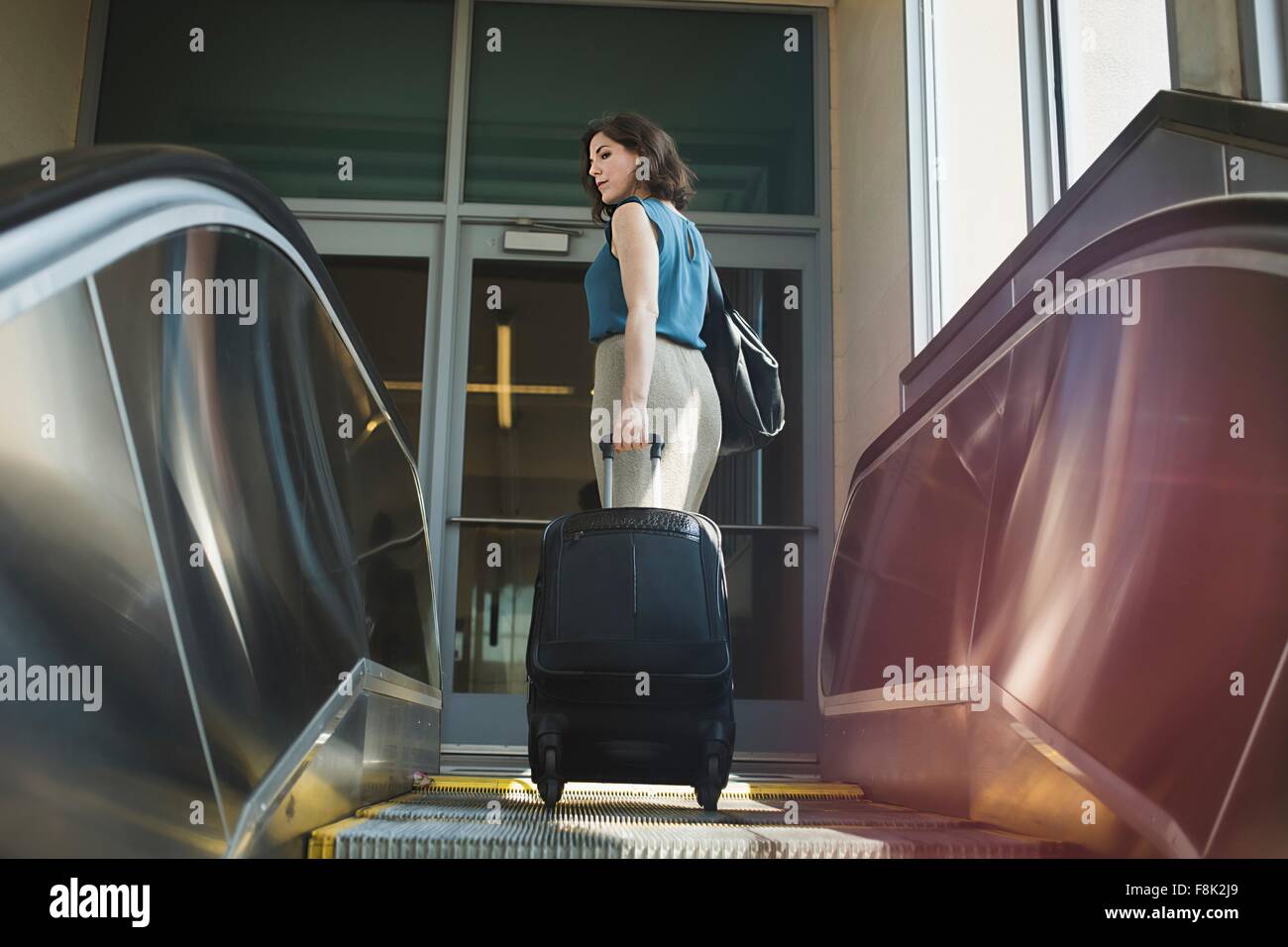 Mid adult woman using escalator, holding wheeled suitcase, rear view Stock Photo