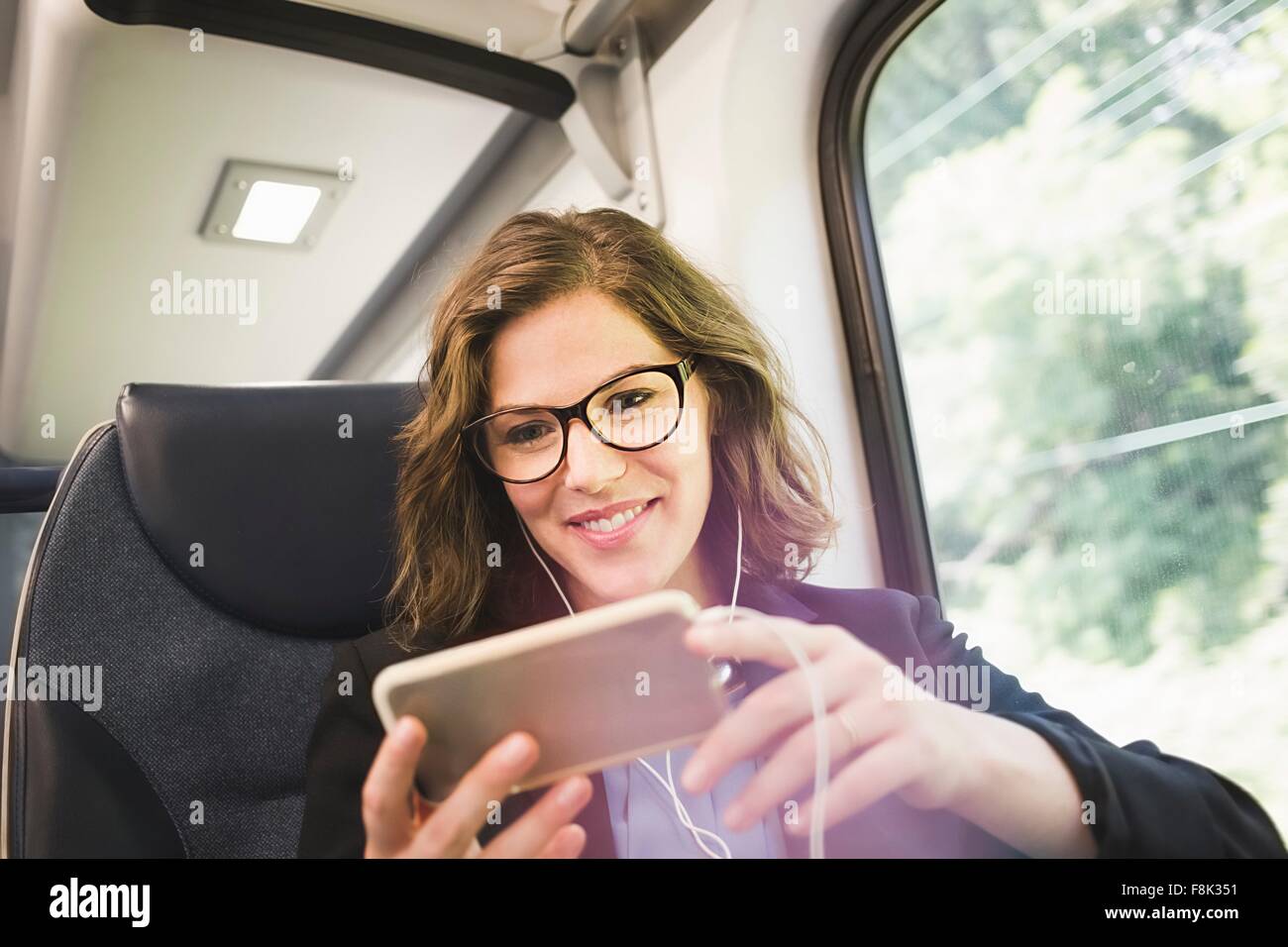 Mid adult woman on train, using smartphone, wearing earphones Stock Photo