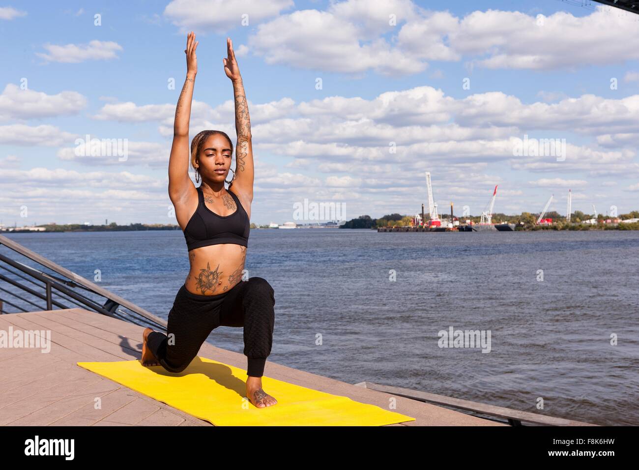 Young woman by water on one knee arms raised in yoga position, eyes closed, Philadelphia, Pennsylvania, USA Stock Photo