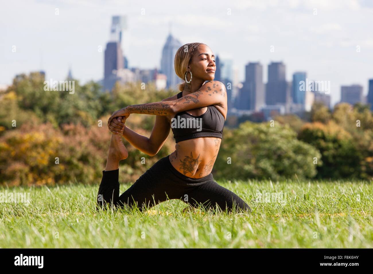 Front view of young woman kneeling on grass stretching leg in yoga position, looking away, Philadelphia, Pennsylvania, USA Stock Photo