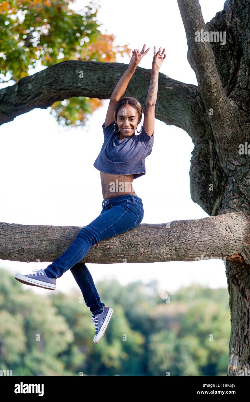Young woman sitting in tree arms raised looking at camera smiling Stock Photo