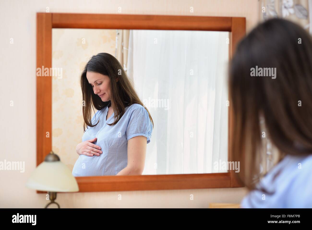 Pregnant woman holding stomach, standing in front of mirror Stock Photo