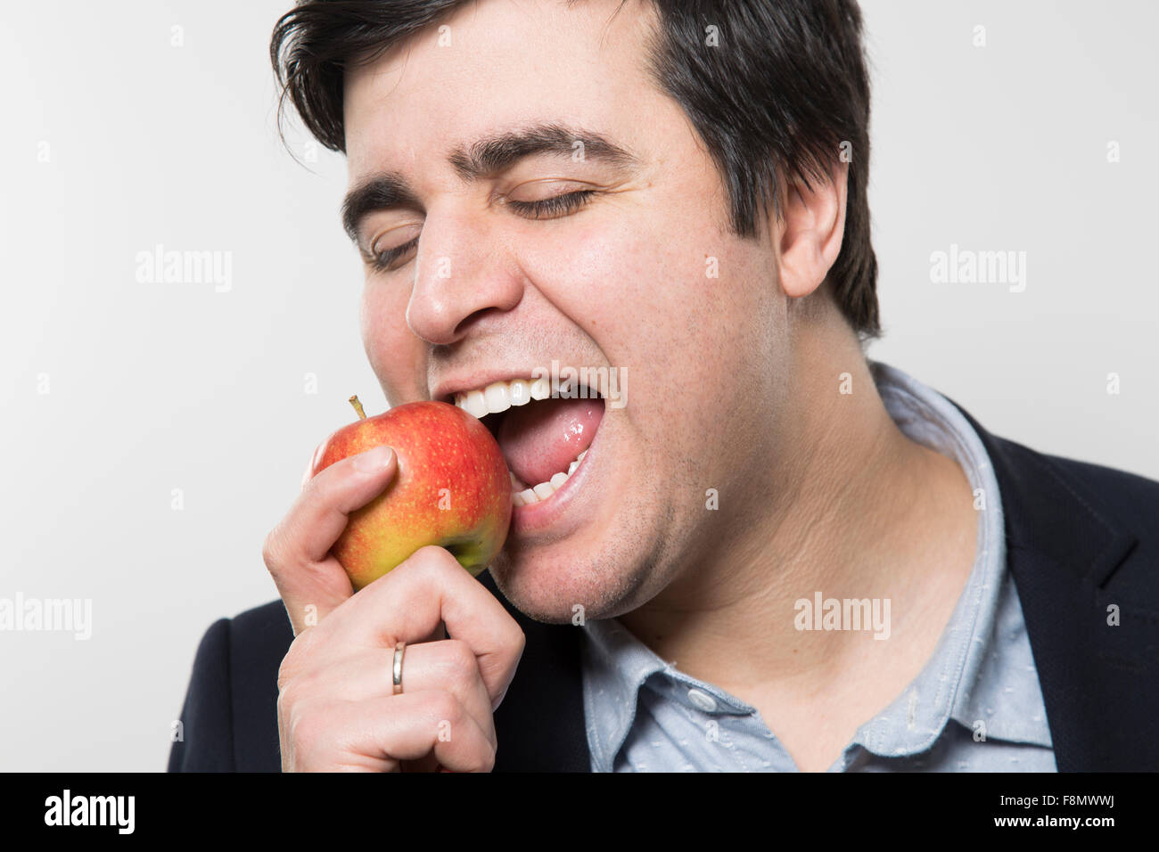 Dark-haired european businessman with a cheerful look takes a bite out of an small apple with his eyes closed while in front of  Stock Photo