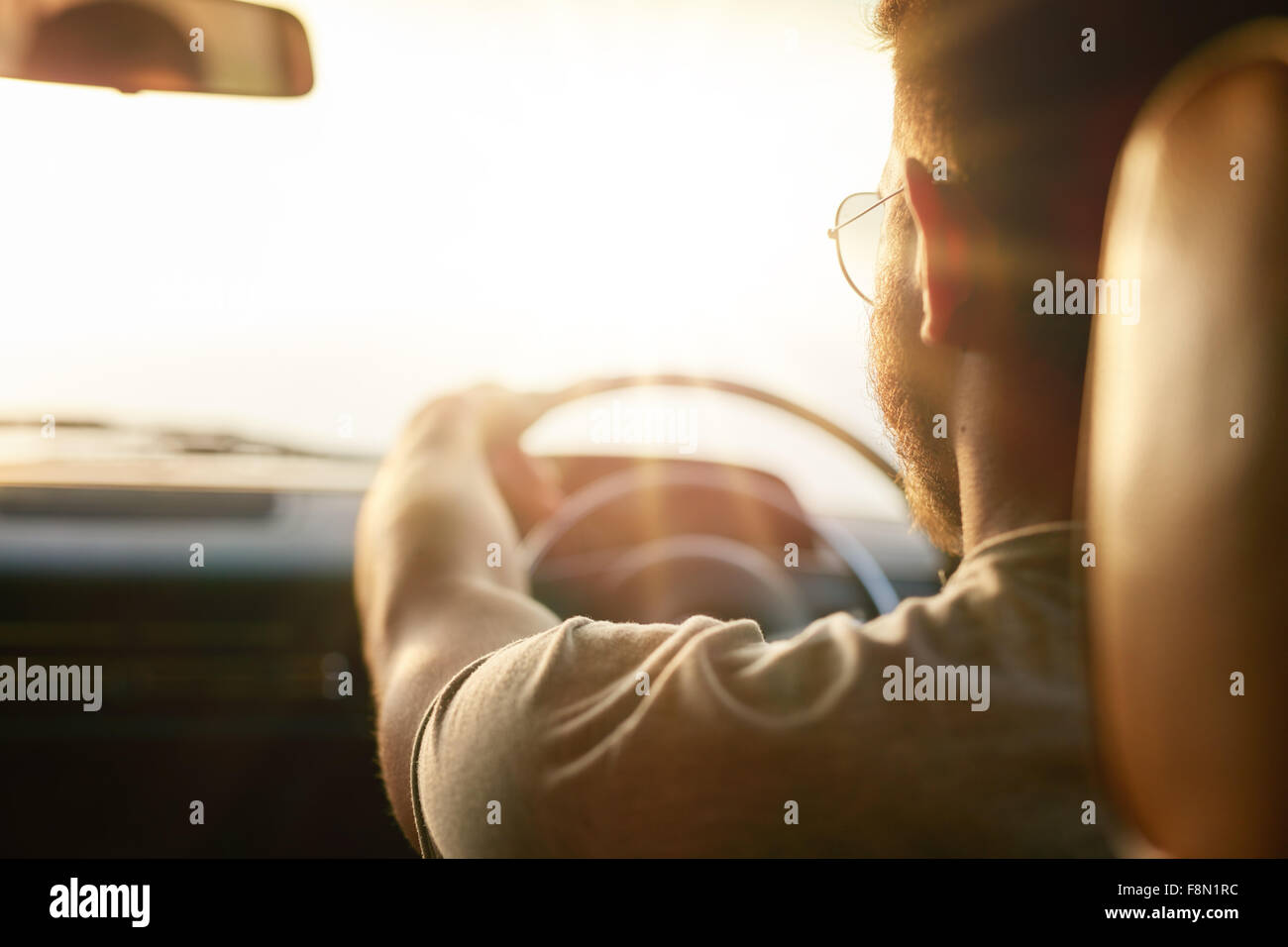 Close up shot of young man driving car. Rear view of man on road trip on a summer day, with sun flare. Stock Photo