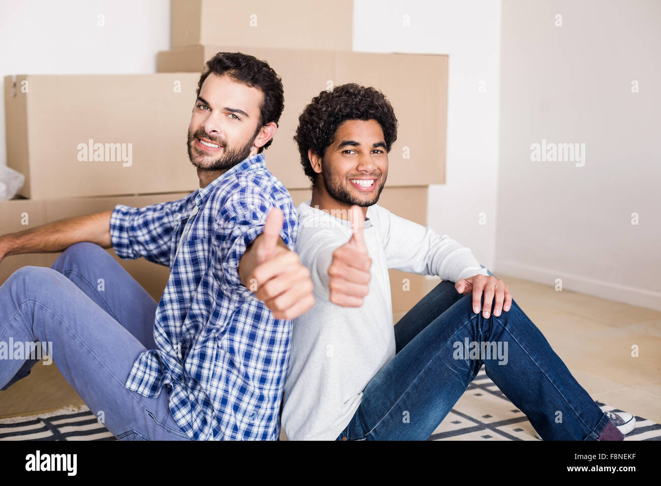 Happy gay couple sitting on floor back-to-back showing thumbs up Stock Photo