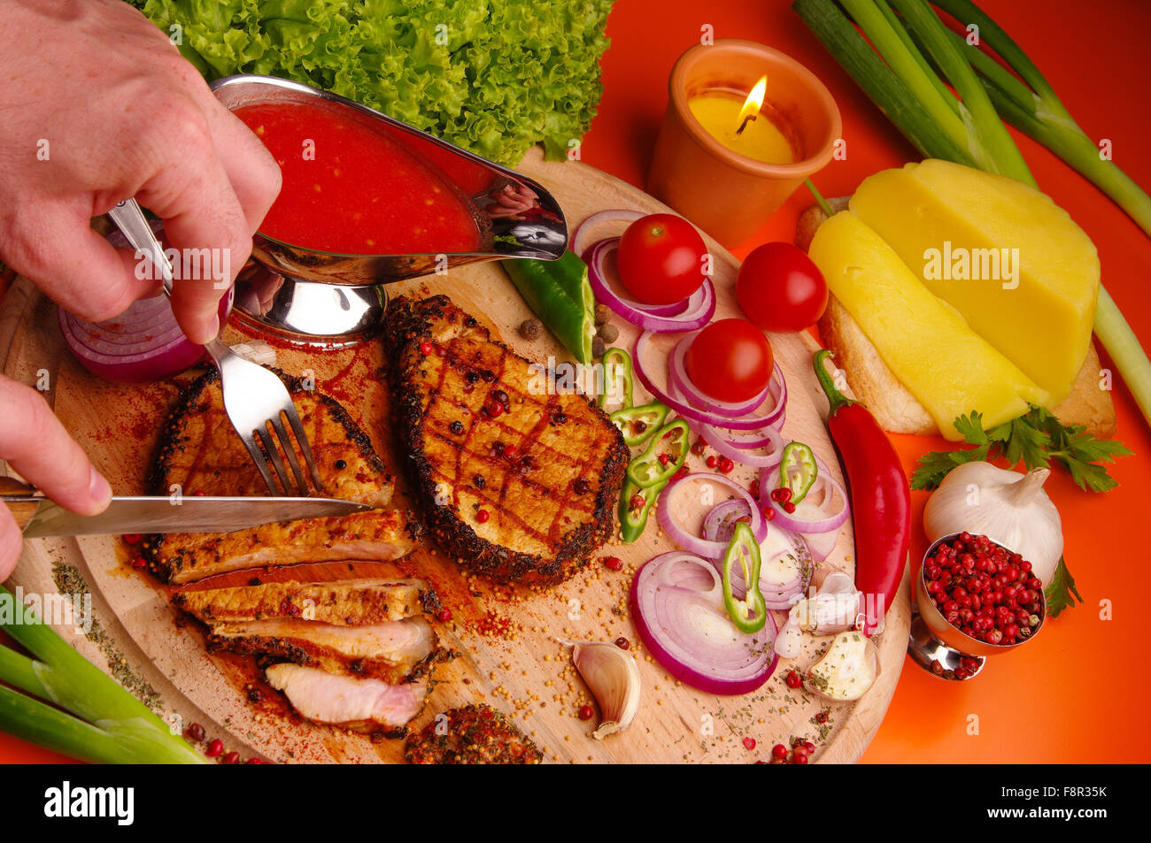 close-up of person cutting a slice of grilled meat, with spices on it and vegetables and cheese around and a silver gravy boat w Stock Photo