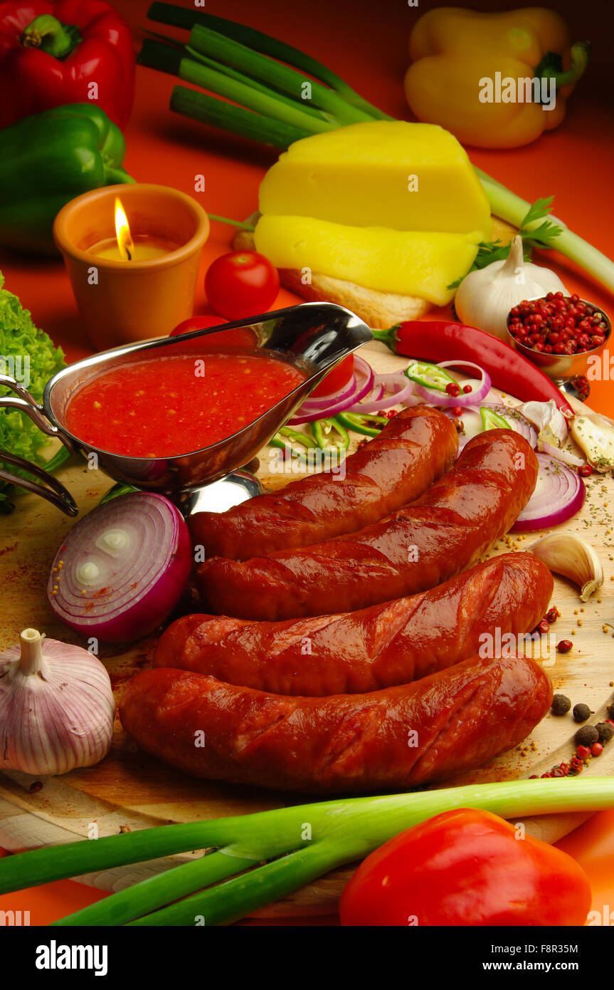 close-up of wooden cutting board with four fried sausages, with spices and vegetables and cheese around and a silver gravy boat Stock Photo