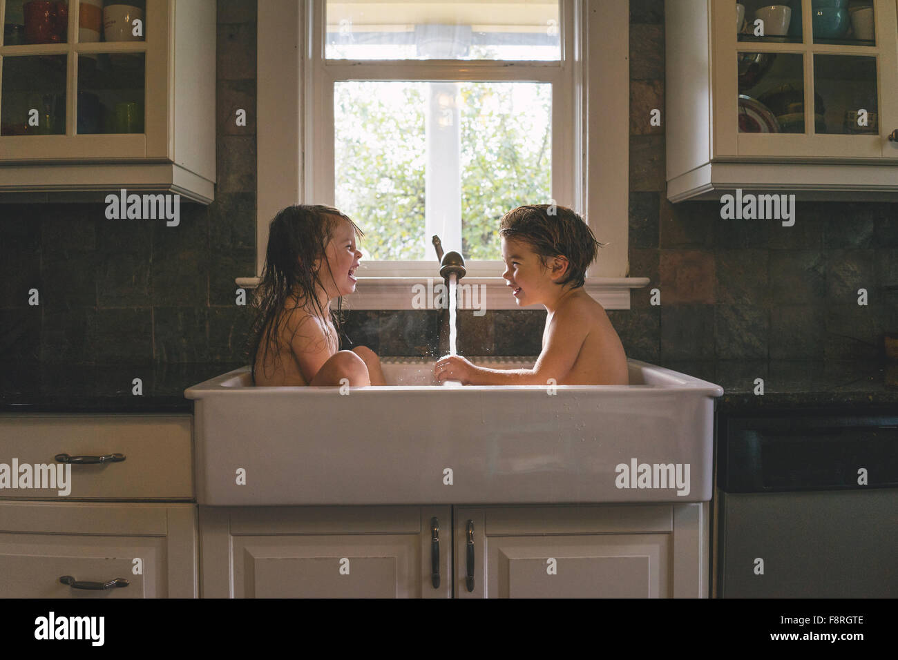 Young boy and girl sitting in kitchen sink playing with water Stock Photo