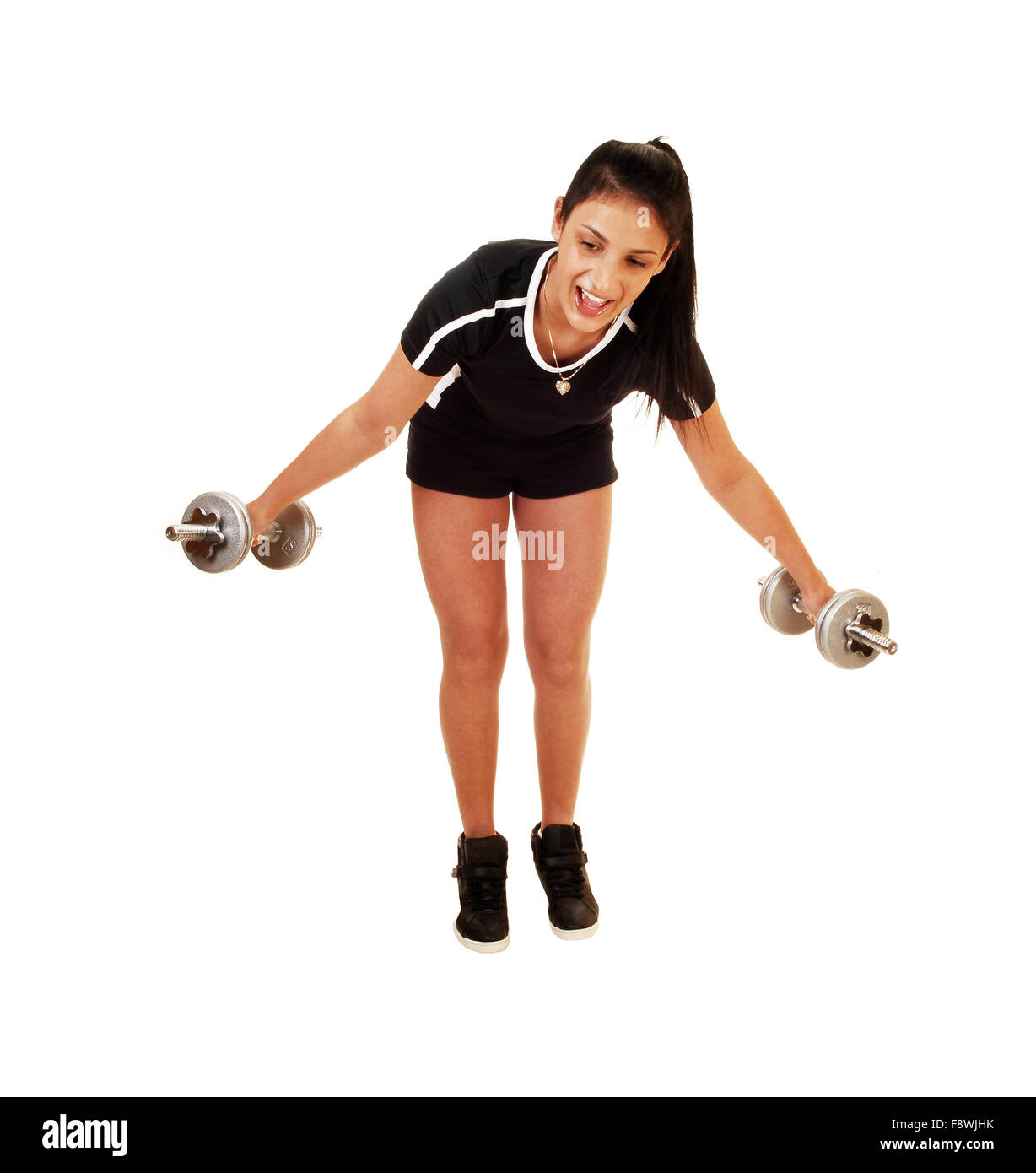 A teenager girl in a black top and shorts standing for white background lifting two dumbbells to exercise her body. Stock Photo