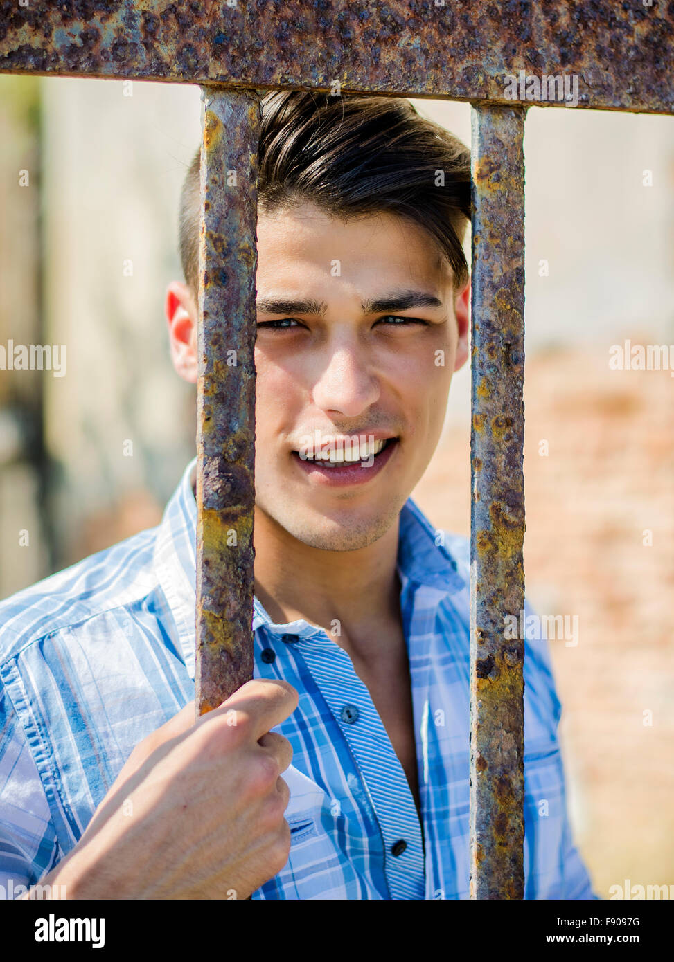 Handsome smiling black haired, blue eyed young man in shirt behind metal cage or gate bars Stock Photo