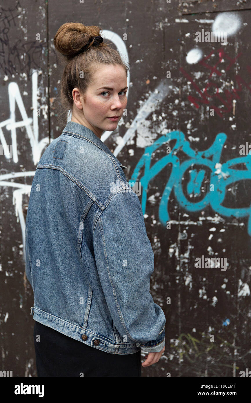 Cardiff, Wales, UK, November 09, 2015. Girl in a blue jeans jacket in front of brown wall with graffities looking over her shoul Stock Photo