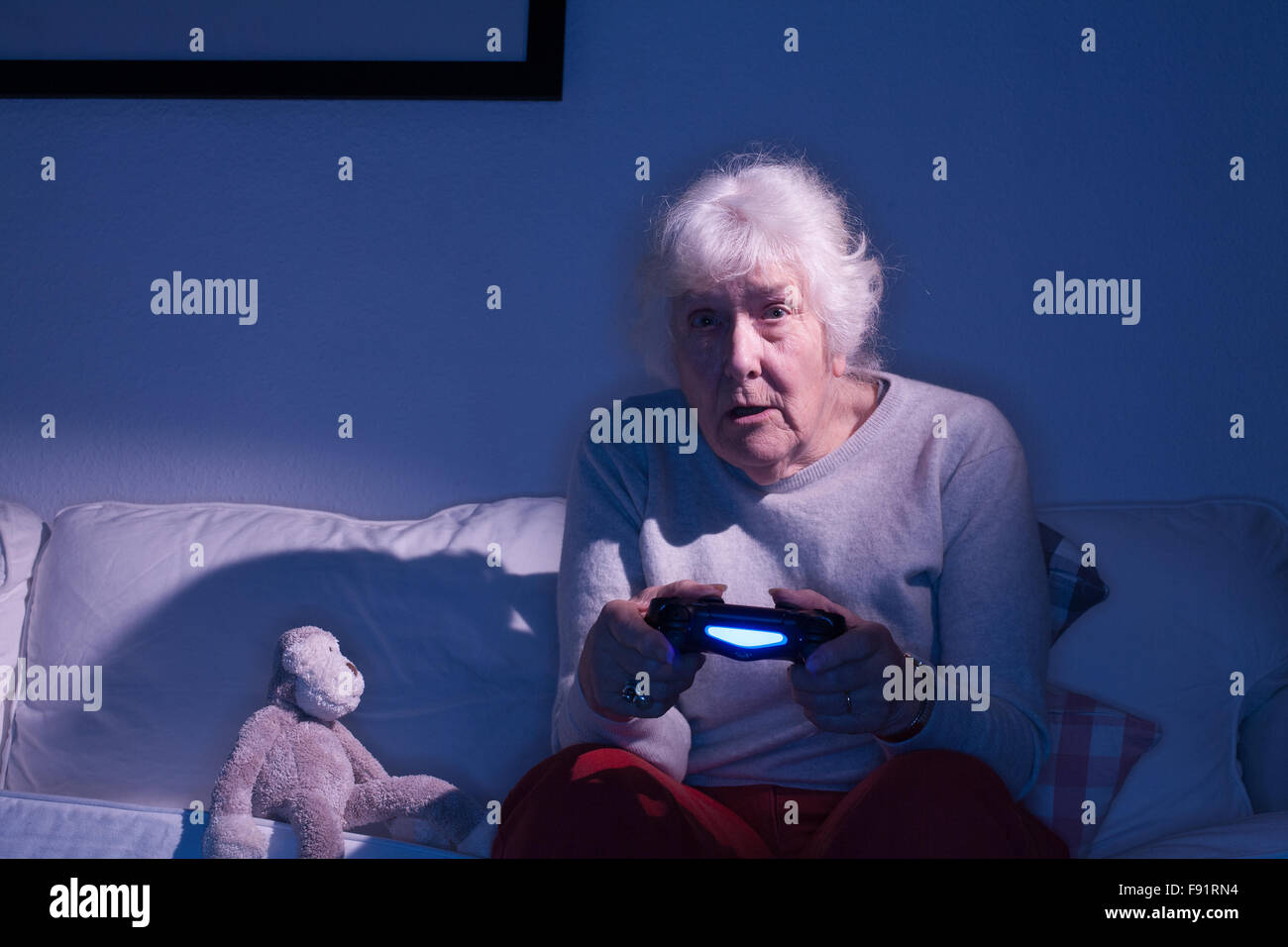 Elderly lady sitting on a sofa playing on a Playstation 4 next to a stuffed toy monkey. Stock Photo