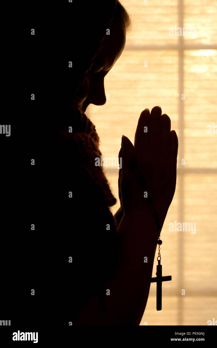 Woman praying silhouetted by window light holding a rosary Stock Photo ...
