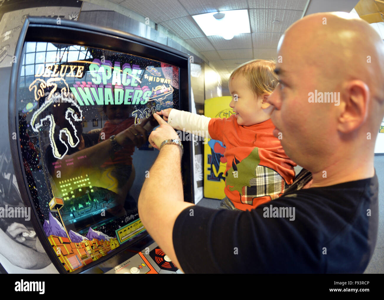 New York, USA. 12th Dec, 2015. DANNY TORRES holds his 19-month-old son MATEO TORRES, from Lindenhurst, as they point to yeti-like aliens on the Midway Bally 1980 video game Deluxe Space Invaders, which the dad played during Opening Day of Arcade Age exhibit, at Cradle of Aviation Museum in Long Island. Admission includes unlimited free pay-to-play of video arcade games, and games history is displayed outside arcade area. Exhibit runs from Dec. 12, 2015 through April 3, 2016. © Ann Parry/ZUMA Wire/Alamy Live News Stock Photo