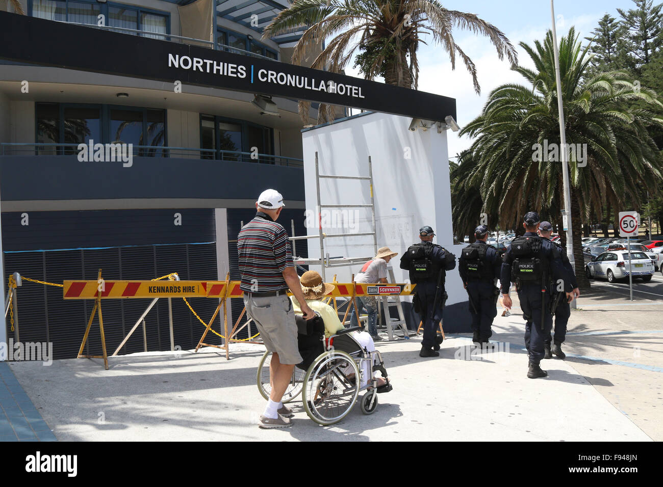 An elderly man pushes an elderly woman in a wheelchair past Northies Cronulla Hotel as NSW police riot squad patrol Cronulla. Stock Photo