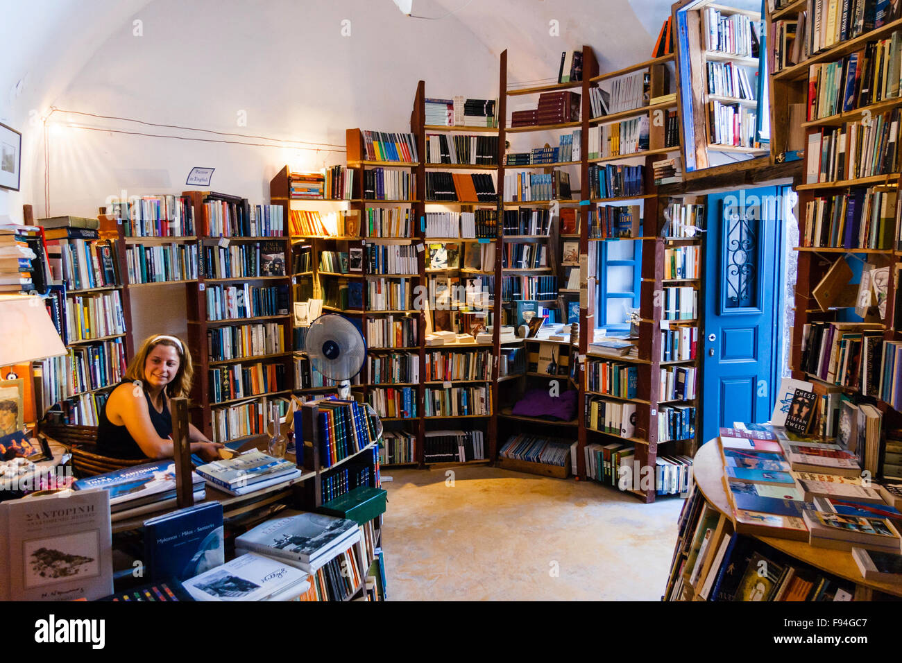 Santorini, Oia. Atlantis second hand bookshop. Interior,  walls covered in wooden shelves full of books, woman shop assistant sitting at desk, smiling. Stock Photo