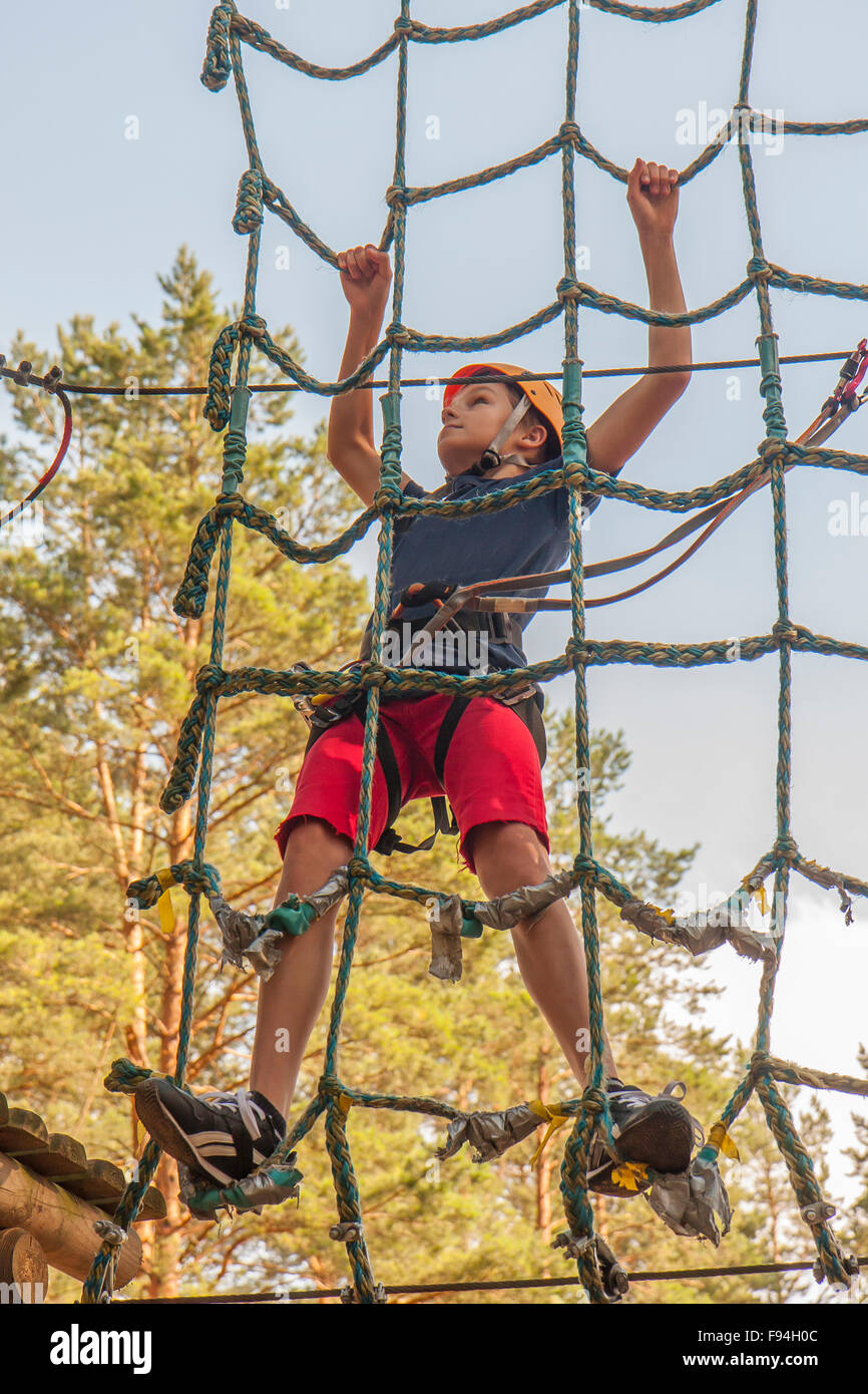 A boy wearing a safety harness and a helmet walking in a rope park ...