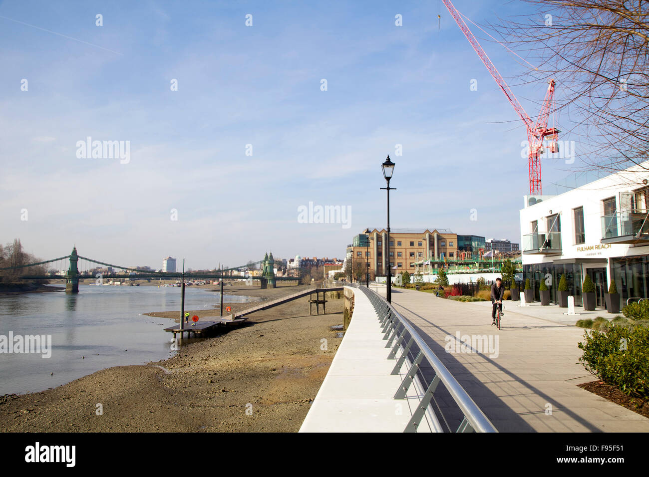 Hammersmith, London. View of the Thames Path with a cyclist riding on it. Building crane and bridge in the distance. Stock Photo