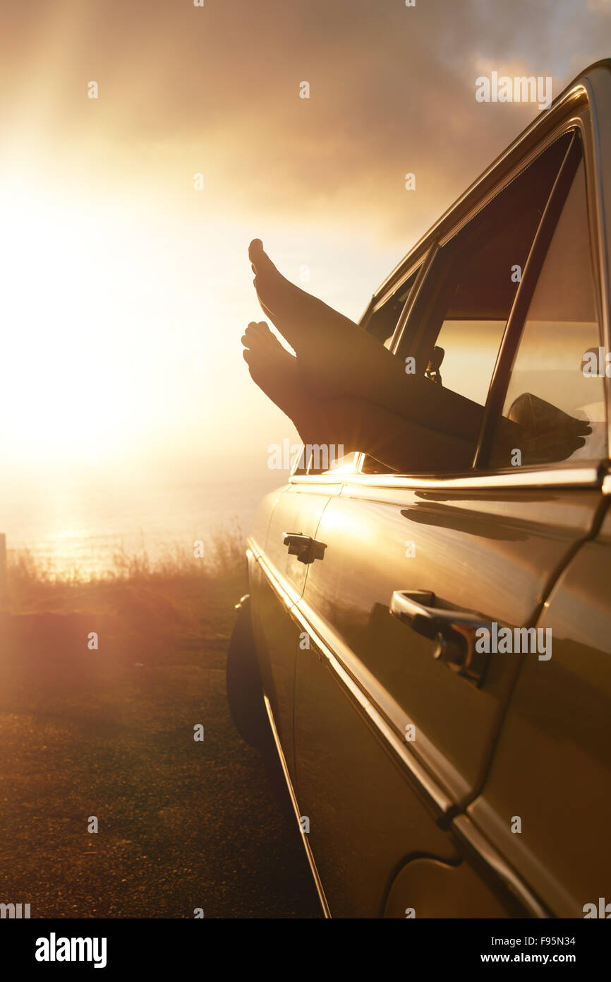 Summer holidays road trip concept. Woman hanging her legs out of car window  at sunset. Stock Photo