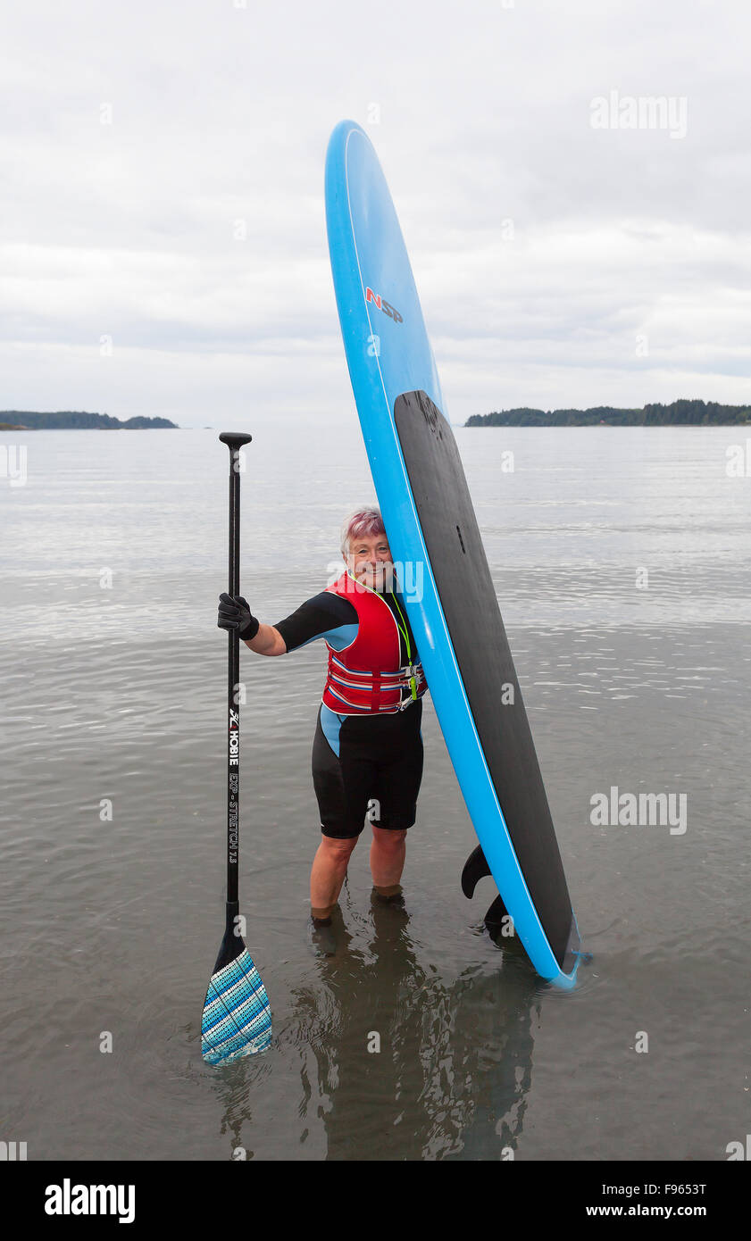 A senior woman comes ashore with her stand up paddle board after paddling in the waters off of Stories Beach near Port Hardy Stock Photo