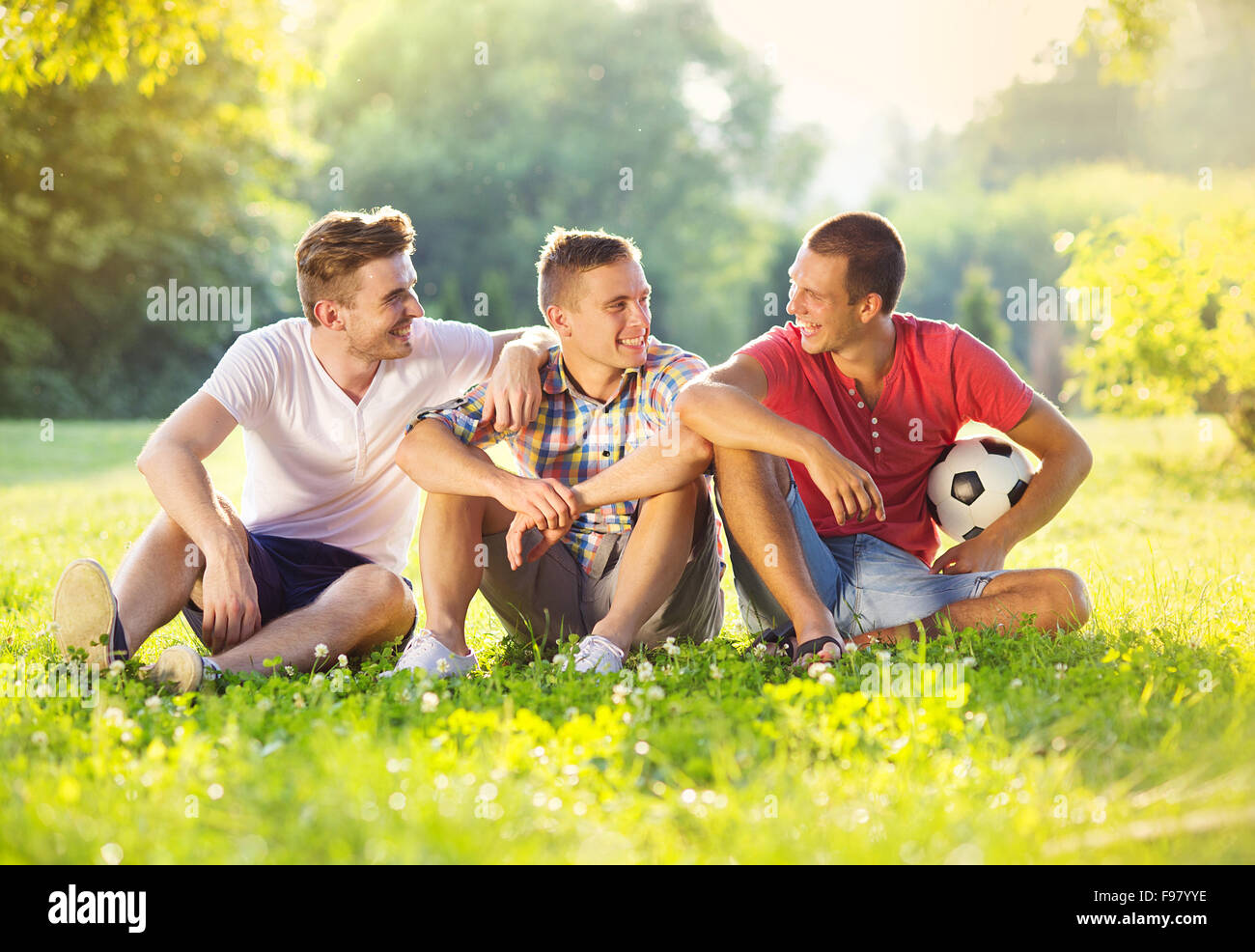 Three happy friends spending free time together in park sitting on grass and chatting Stock Photo