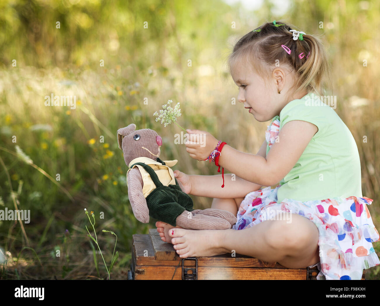 Summer outdoor portrait of cute little girl sitting on meadow with old suitcase with toys Stock Photo