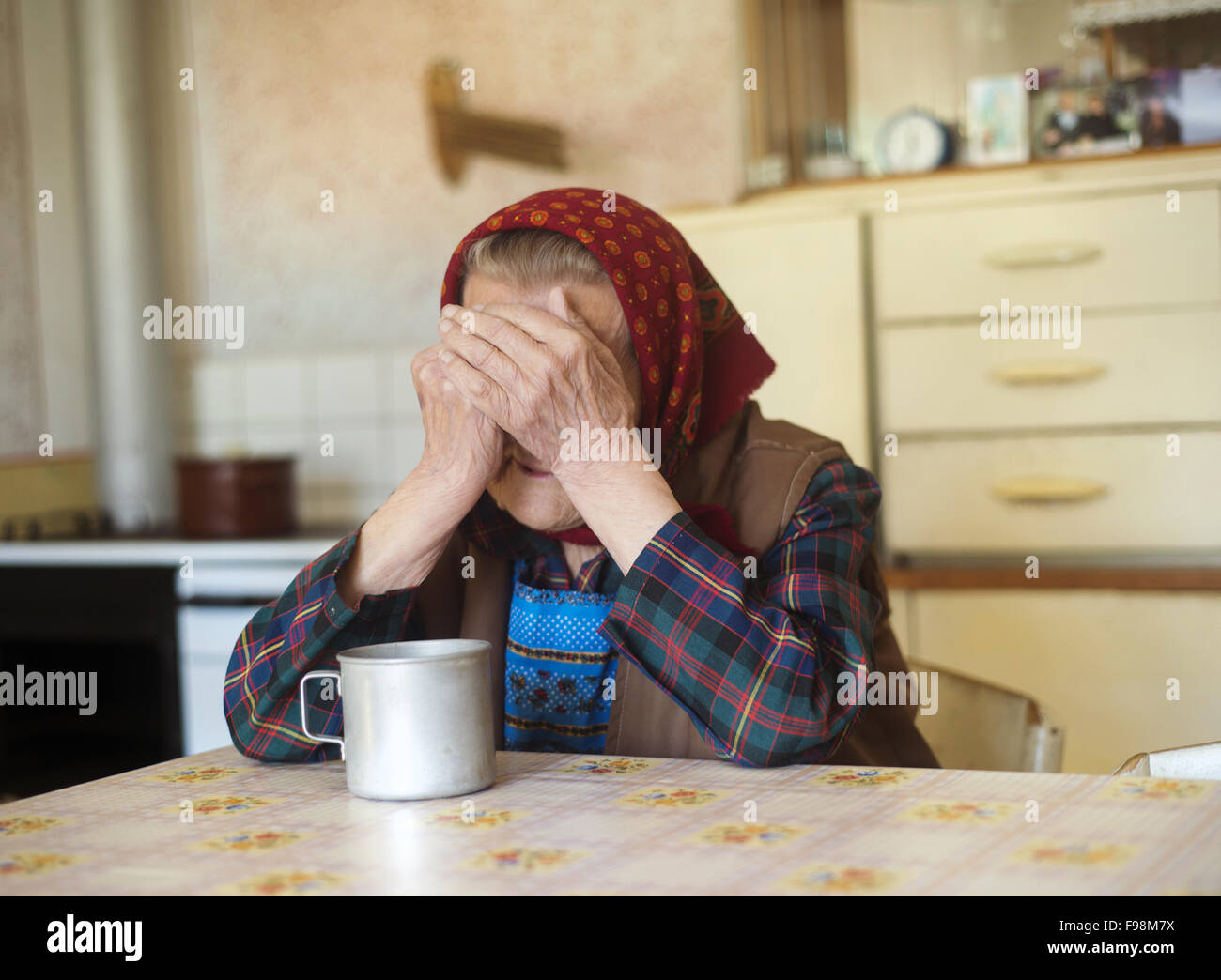 Very old sad woman in headscarf sitting in her countrystyle kitchen Stock Photo