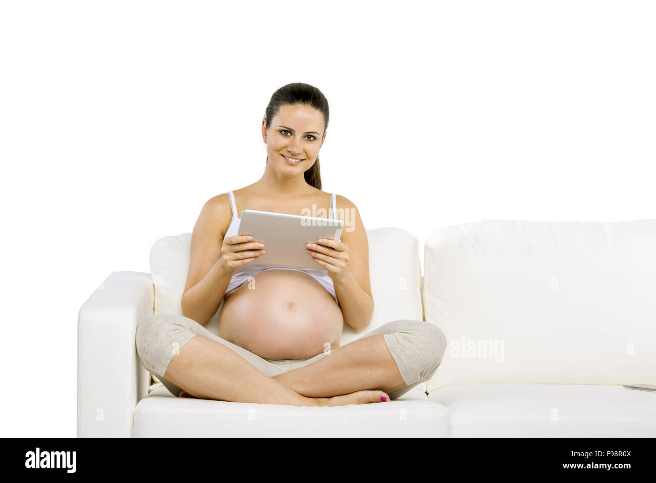 Studio portrait of pregnant woman with digital tablet sitting on sofa isolated on white background Stock Photo