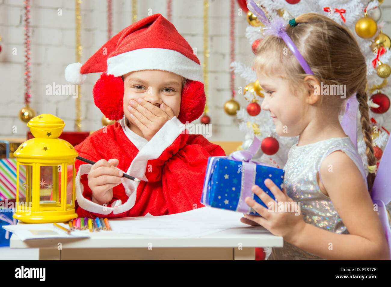A girl dressed in a red suit Santa Claus draws pencils sitting at the table, next is a girl dressed as a fairy with the candlest Stock Photo
